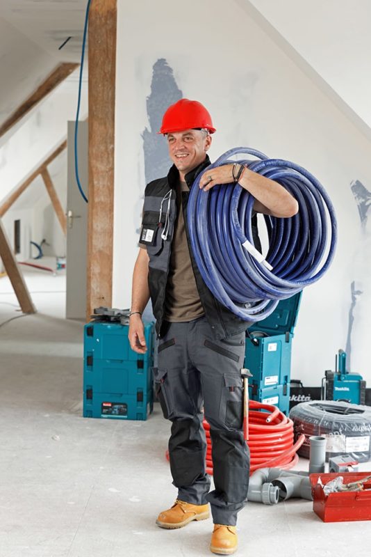 Homme sur un chantier ayant enrouler sur une épaule des gaines électriques bleues. il porte un casque de chantier et des chaussures de sécurité vendu par abisafe